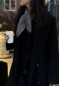 Manteau noir avec grands revers, écharpe grise, tenant une tasse blanche. Vêtements sombres, texture lisse. En arrière-plan, des plantes d'intérieur et du bois clair.