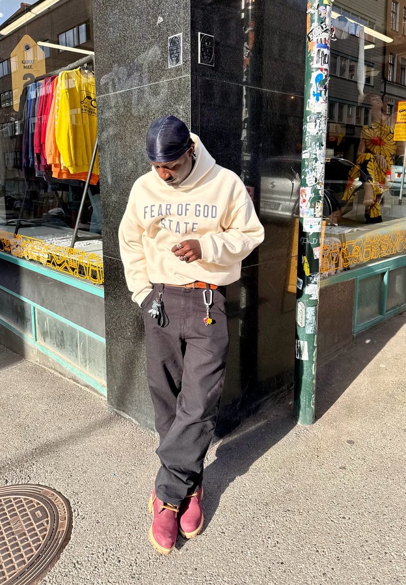 Cream hoodie with "FEAR OF GOD STATE" text, black pants, and burgundy boots. Keychains attached to pants. Smooth fabric and casual style.