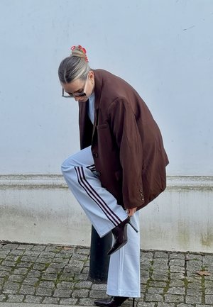 Mujer con gafas de sol y pinza en el cabello ajusta una bota negra de tacón alto, vistiendo un blazer marrón oversize y pantalones blancos con rayas moradas.