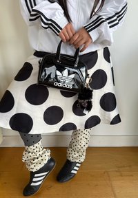 Černá kabelka z lakované kůže s bílým logem Adidas, bílá puntíkovaná sukně, šedé legíny s puntíky a černé boty Adidas s bílými pruhy.
