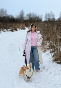 Femme en pull rose et manteau blanc promenant un chien corgi sur un chemin enneigé avec de l'herbe sèche et des arbres en arrière-plan.