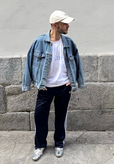 Chaqueta de mezclilla sobre una camiseta blanca, pantalones de chándal azul marino con rayas laterales y zapatillas plateadas, todo exhibido contra un fondo de pared de piedra.