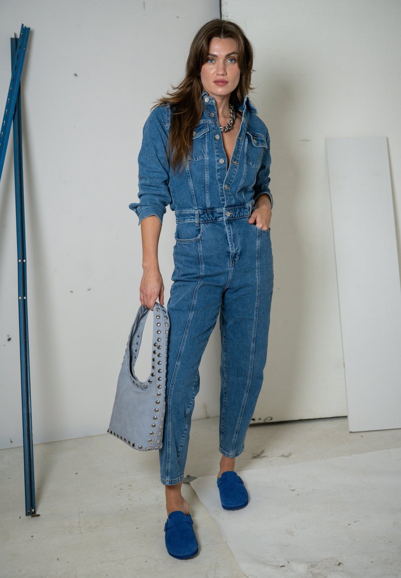 Donna che indossa una tuta di jeans aderente, tiene in una mano una borsa grigia con borchie e indossa scarpe slip-on blu, in piedi all'interno di un edificio.