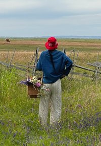 Kosz wypełniony kolorowymi kwiatami; osoba w niebieskim swetrze i w paskowanych spodniach, nosząca czerwoną kapelusz typu bucket, stoi na trawiastym polu.