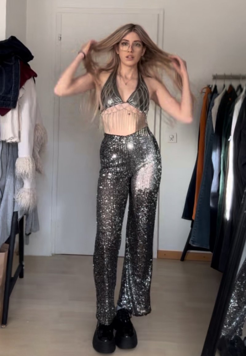 Srebrny top na szyję z cekinami i frędzlami, połączony ze srebrnymi, szerokimi spodniami zdobionymi cekinami. Noszony z czarnymi butami na platformie.