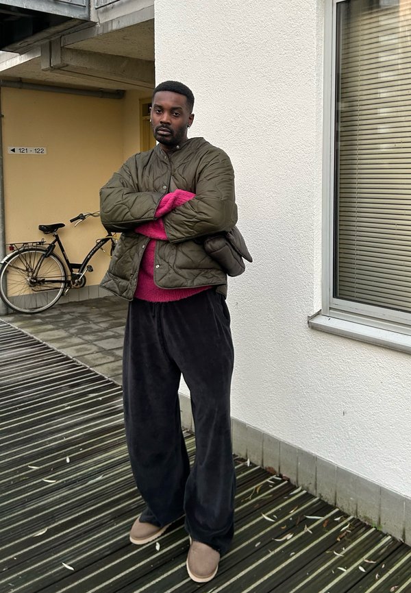 Veste matelassée verte, pull rose, pantalon large noir, chaussures marron ; debout sur des planches de bois près d'un mur et d'un vélo.
