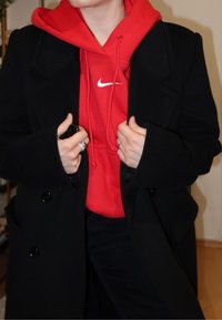 Manteau en laine noire porté sur un sweat à capuche rouge avec logo blanc, présentant de gros boutons, de larges revers et une coupe décontractée. Mains ornées de bagues visibles.