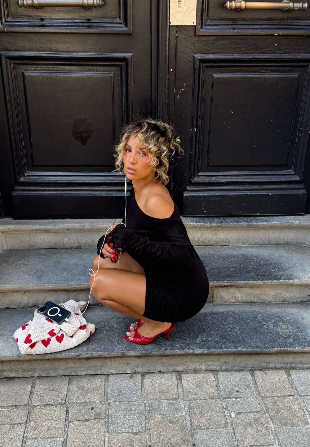 Svart off-shoulder kjole med tettsittende silhuett. Røde høye hæler med sløyfedetalj. Hvit veske med rødt hjerte mønster og en smarttelefon. Grå steintrinn.