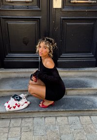 Svart off-shoulder kjole med tettsittende silhuett. Røde høye hæler med sløyfedetalj. Hvit veske med rødt hjerte mønster og en smarttelefon. Grå steintrinn.