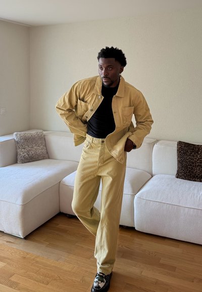 Hombre ajustándose chaqueta y pantalones beige, con camisa negra y zapatos estampados, de pie junto a un sofá seccional blanco con cojines texturizados.