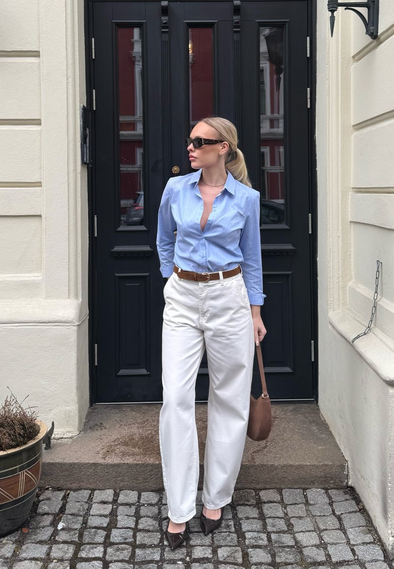 Femme portant une chemise bleu clair, un pantalon blanc, une ceinture marron et des lunettes de soleil, se tenant sur un trottoir en pavés devant une porte noire.