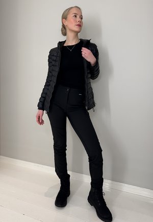 Chaqueta de abrigo negra con cremallera, camiseta negra de manga larga, pantalones negros ajustados y botas negras, sobre un fondo neutro.