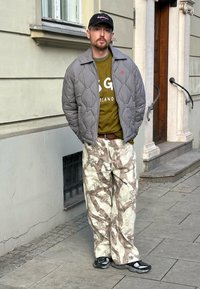 Jaqueta bomber cinza matelassada sobre uma t-shirt de logo verde-oliva, combinada com calças largas em creme e castanho com padrão e sandálias pretas.