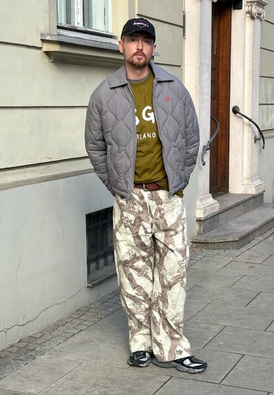 Chaqueta bomber acolchada gris sobre una camiseta con logo verde oliva, combinada con pantalones holgados crema y marrón con patrón y sandalias negras.