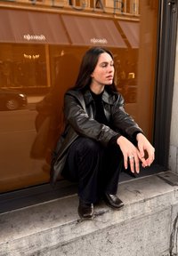 Melnas ādas jaka, melns tops un melnas bikses. Brūni ādas zābaki. Persona sēž uz akmens pakāpieniem pie oranžas spoguļainas loga.