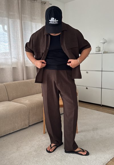 Camisa de botones de manga corta marrón sobre una camiseta sin mangas negra, combinada con pantalones anchos marrones y chanclas negras; diseño y textura sencillos.