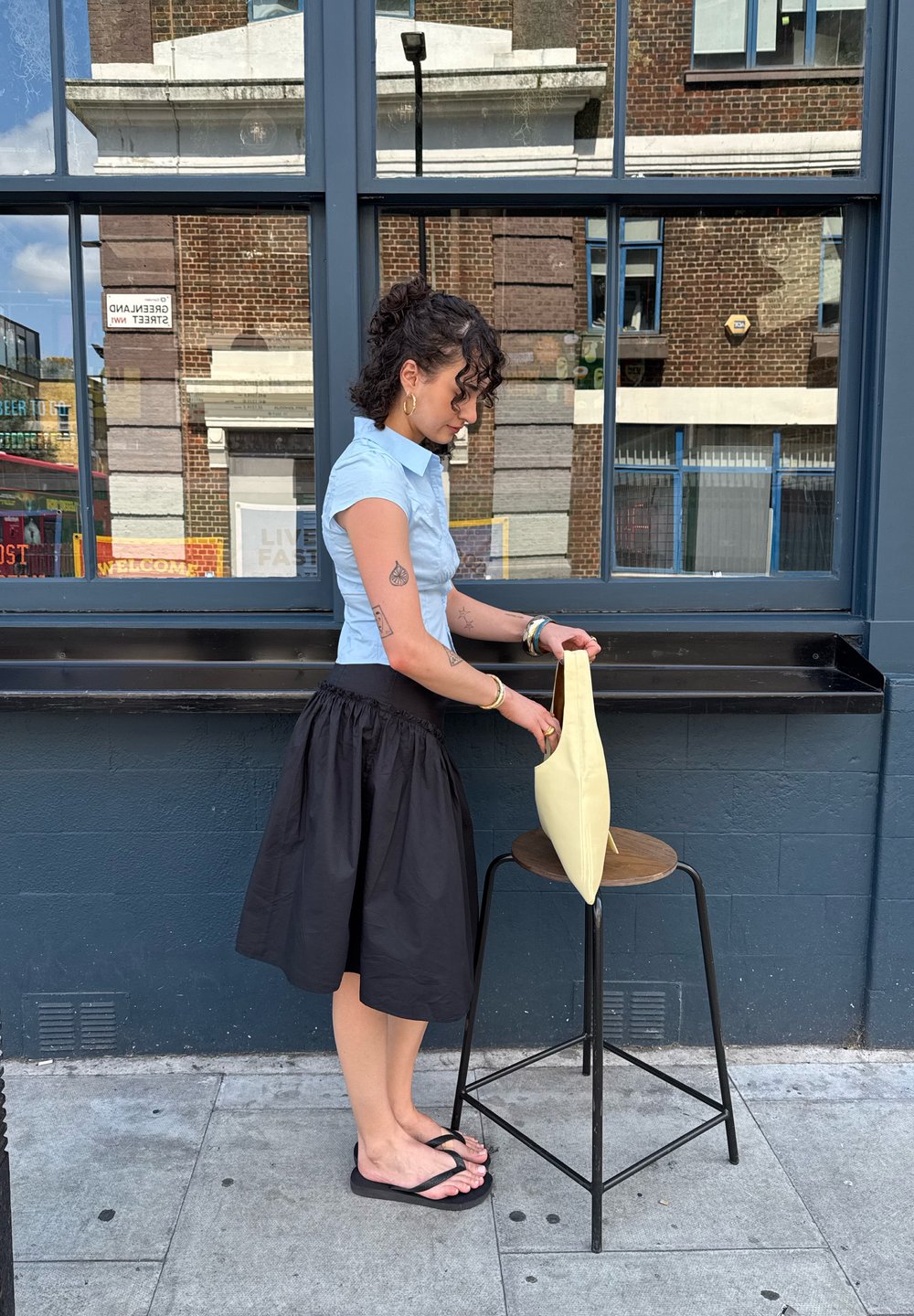 Borsa gialla a forma conica esposta su uno sgabello di legno accanto a una persona con camicia azzurro chiaro e gonna nera, in piedi sul marciapiede.