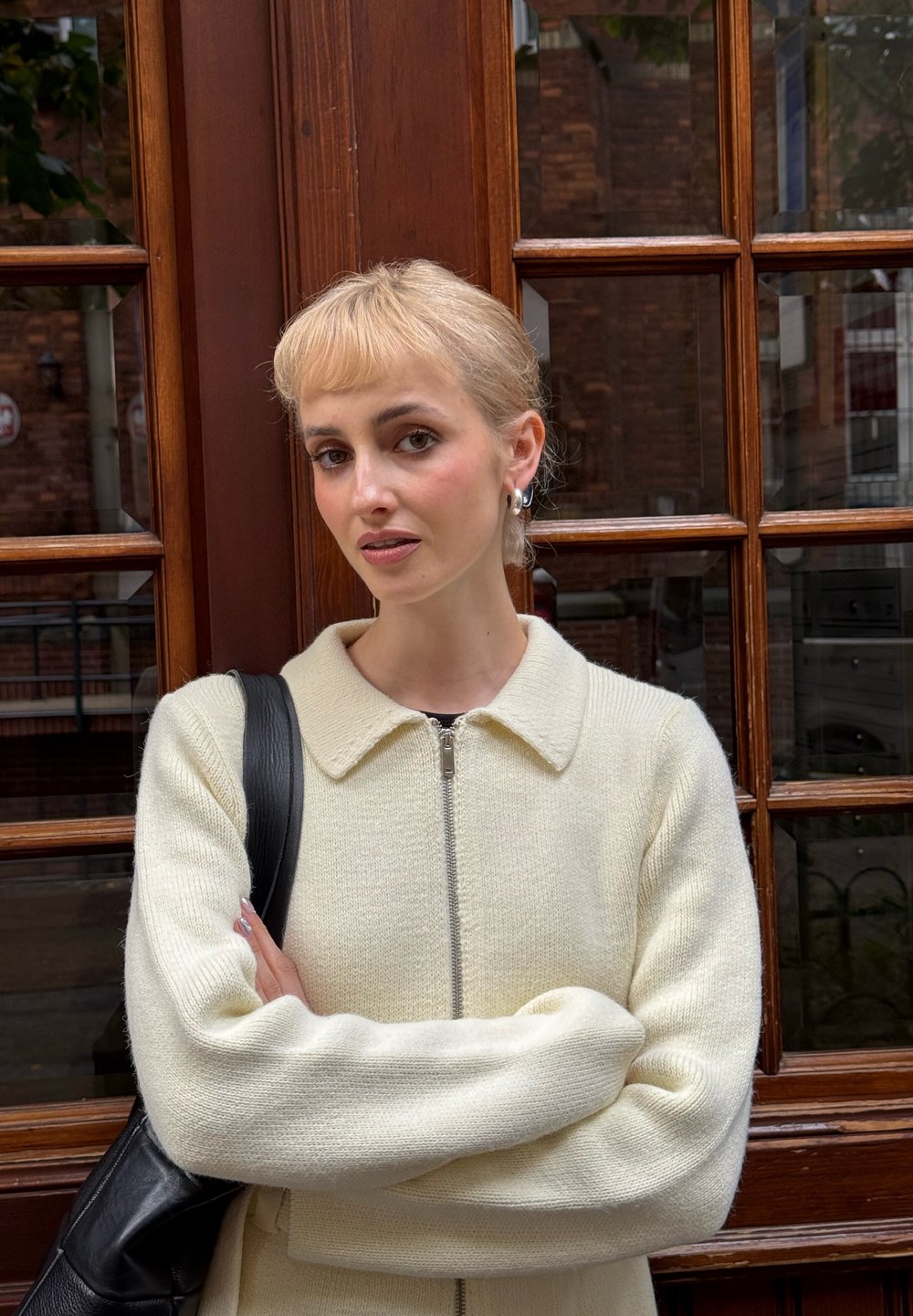 Suéter de punto color crema con cuello y cremallera. La modelo lleva un bolso negro de hombro, apoyada contra un marco de ventana de madera.