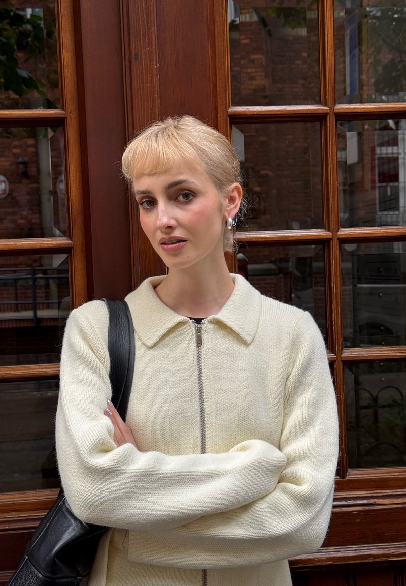 Cremefarbener Strickpullover mit Kragen und Reißverschluss. Modell trägt eine schwarze Schultertasche und steht vor einem hölzernen Fensterrahmen.