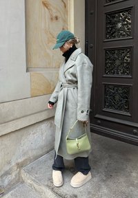 Grau wollener Wickelmantel mit einem Gürtel, schwarzem Rollkragenpullover, grünem Hut, grüner Handtasche und beigen Slip-On-Schuhen, vor einer strukturierten Wand.