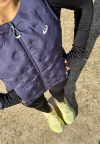 Granatowa kamizelka puchowa z okrągłymi wzorami, z zamkiem z przodu, noszona na czarnej bluzce z długim rękawem, w zestawie z czarnymi legginsami i żółtymi sneakersami.