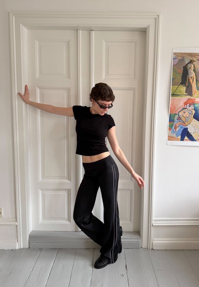 Persona con ropa negra y gafas de sol posa con un brazo apoyado en una puerta blanca con paneles y el otro extendido hacia un lado en una habitación luminosa.