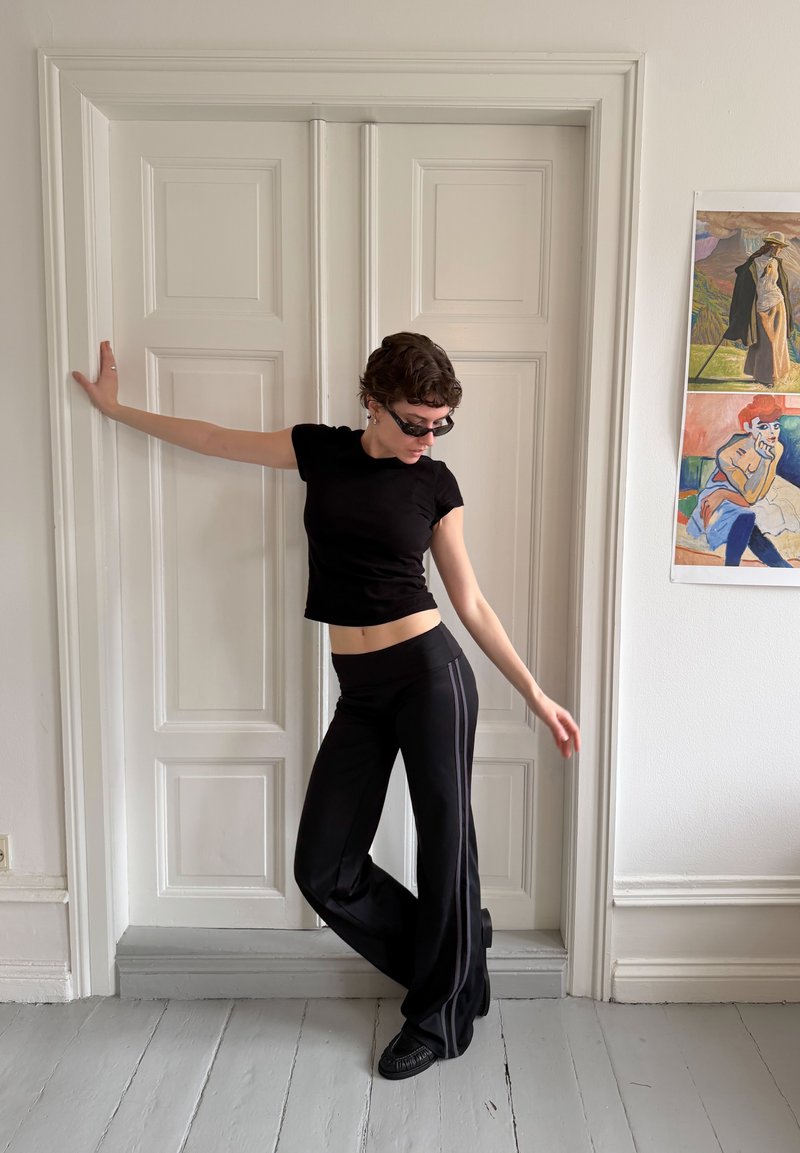 Personne portant une tenue noire et des lunettes de soleil pose avec un bras sur une porte à panneaux blanche et l'autre bras étendu sur le côté dans une pièce lumineuse.