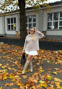 Gestreepte beige gebreide trui, korte witte rok, beige laarzen tot de knie, zwarte handtas met studs, herfstbladeren op de grond.