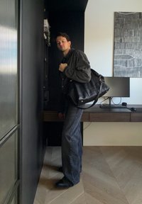 Homme portant une chemise à carreaux et un jean, portant un grand sac en cuir noir dans une pièce moderne avec un bureau, un ordinateur et une œuvre d'art murale abstraite.