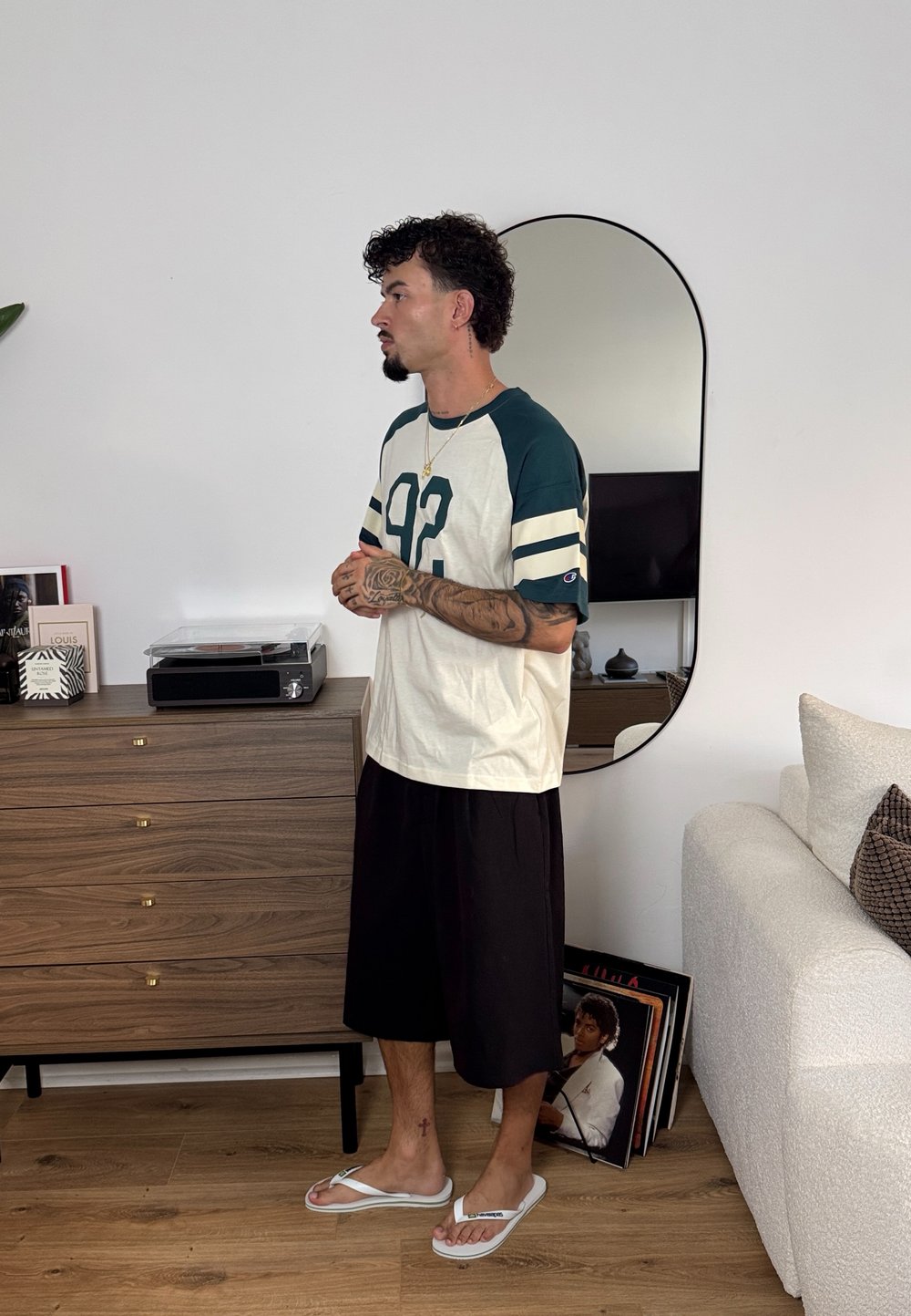 Camicetta bianca e verde con grafica "92", pantaloni corti neri fino al ginocchio e infradito bianche. Sullo sfondo c'è un comò di legno e uno specchio.