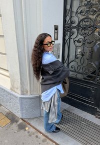 Camisa de mangas longas azul clara sobreposta a um suéter cinza escuro, calças pretas e uma camisa listrada pendurada. De pé ao lado de um portão decorativo.