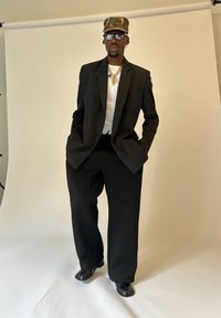 Mann in übergroßem schwarzen Blazer und Hose, weißem Hemd, Camouflage-Cap, Brille und schlichten goldenen Halsketten steht vor einem schlichten Hintergrund.