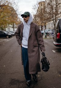 Manteau à carreaux marron, sweat à capuche gris, jean bleu, mocassins noirs et lunettes de soleil. Sac à main noir, décor urbain avec des arbres et des voitures en stationnement.