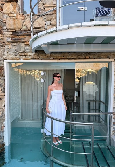Vestido blanco sin tirantes con un corsé ajustado y una falda fluida, de pie en un balcón circular con barandilla de vidrio. Fondo de pared de piedra.