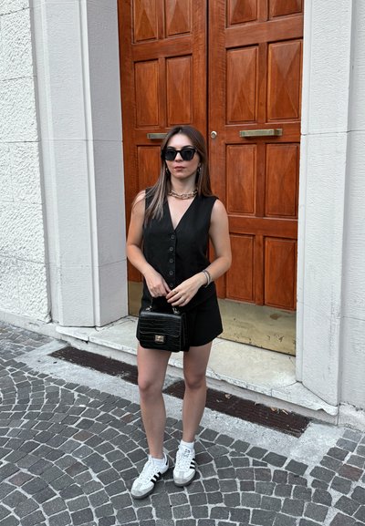 Mono negro sin mangas con detalle de botones, combinado con un collar de cadena gruesa, un bolso negro y zapatillas blancas, con puertas dobles de madera de fondo.