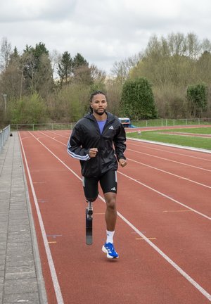 Hombre con una pierna prostética corriendo en una pista de atletismo roja, vestido con una chaqueta negra, pantalones cortos negros y zapatillas de correr azules.