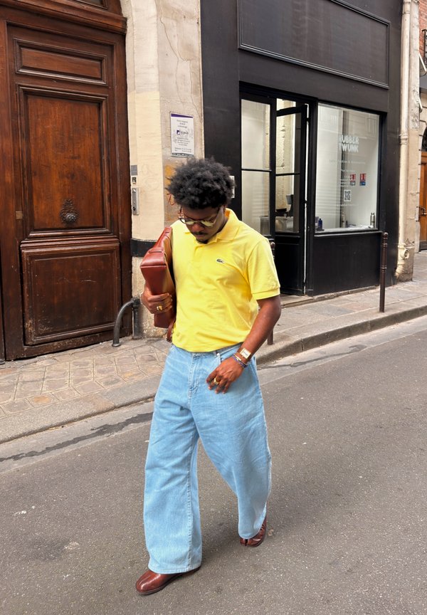 Homme portant un polo jaune et un jean bleu large, marchant dans la rue en tenant un sac en cuir marron sous le bras.