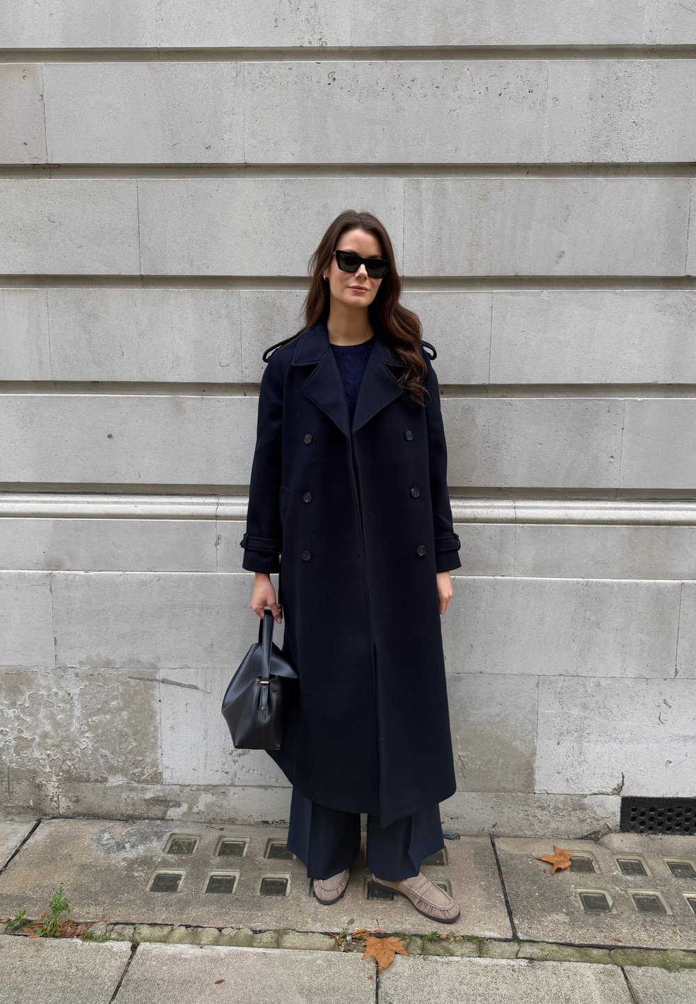 Marineblå dobbelspent frakk i ull med skulderklaffer, kombinert med løse mørke bukser og lysebrune sko med tekstur uten snøring. Svart håndveske.