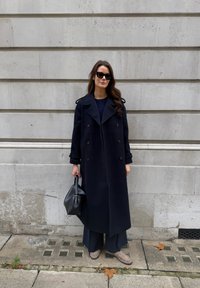 Marineblå dobbelspent frakk i ull med skulderklaffer, kombinert med løse mørke bukser og lysebrune sko med tekstur uten snøring. Svart håndveske.