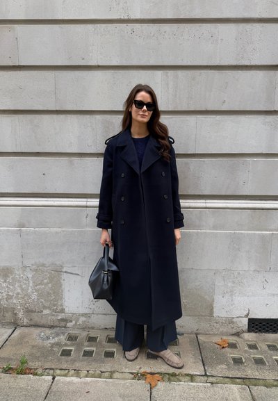 Abrigo azul marino de lana con cierre cruzado y hombreras, combinado con pantalones oscuros y sueltos y zapatos tipo mocasín texturizados de color marrón claro. Bolso negro.