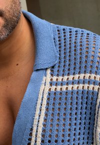 Gaiši zils adīts krekls ar atvērtu, rakstu dizainu un baltām horizontālām svītrām. Iezīmēts ar faktūru un brīvu piegriezumu.