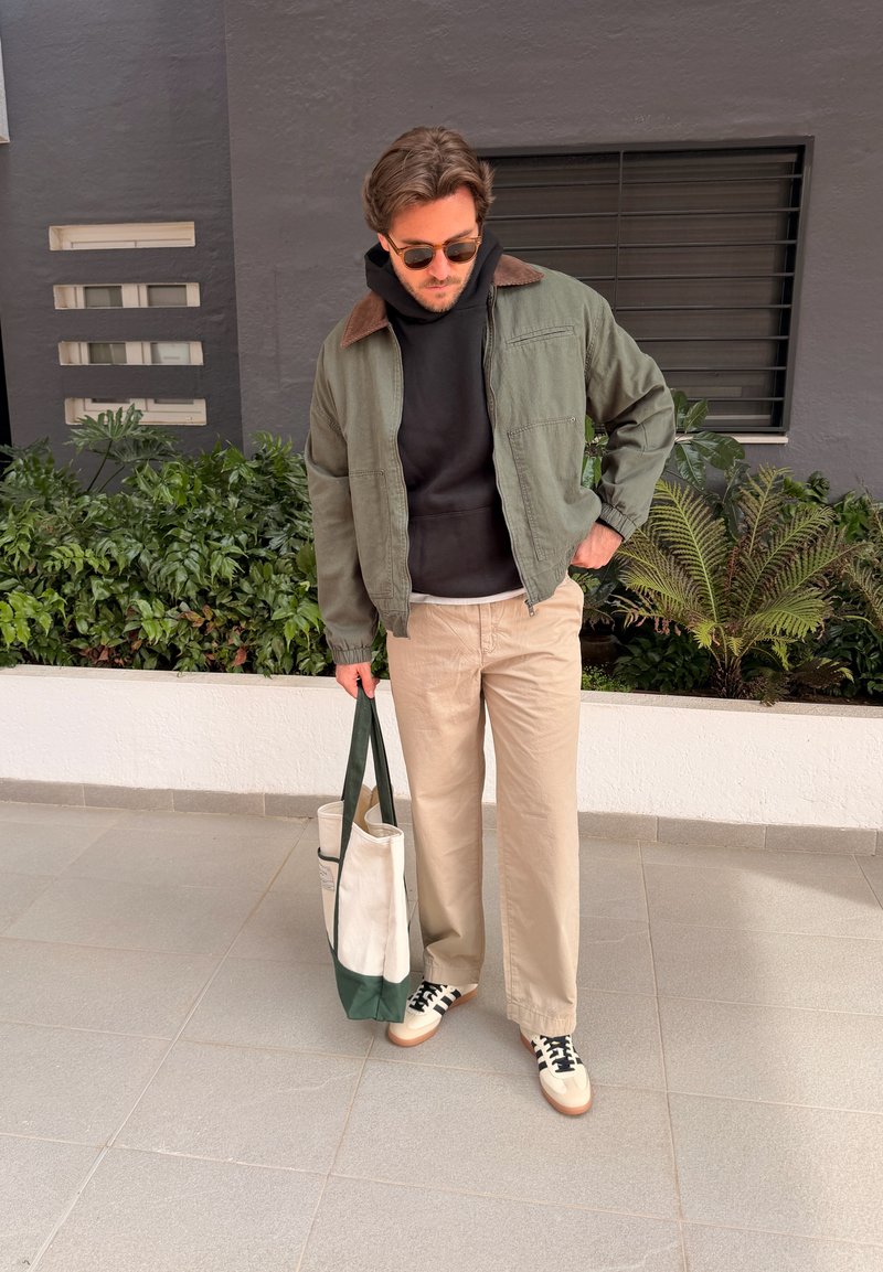 Homme avec lunettes de soleil portant une veste verte, un sweat à capuche noir, un pantalon beige et des baskets, tenant un sac fourre-tout blanc et vert, debout près des plantes.