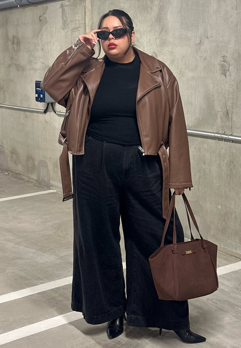 Brun skinnjakke i cropped stil over svart topp, svart vid bukse med høyt liv, svarte hæler og brun håndveske, med solbriller som fremhever antrekket.