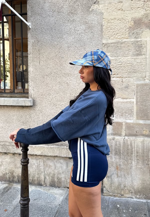 Mujer con una gorra azul a cuadros, pantalones cortos azul marino con rayas blancas y una camiseta azul de manga larga, apoyada en un poste metálico negro junto a una pared de piedra.