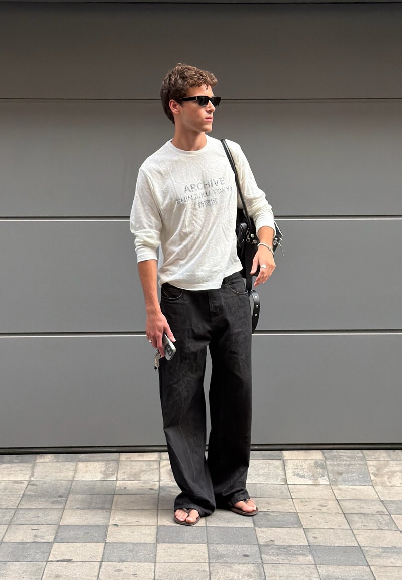 Jeune homme portant des lunettes de soleil, une chemise blanche à manches longues, un pantalon noir et des sandales, debout sur un sol carrelé devant un mur gris tenant un sac.