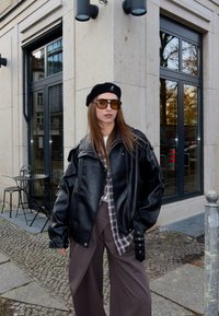 Chaqueta de cuero negra de gran tamaño con solapas grandes, llevada sobre una camisa de cuadros. Pantalones anchos marrones y una boina negra completan el conjunto.