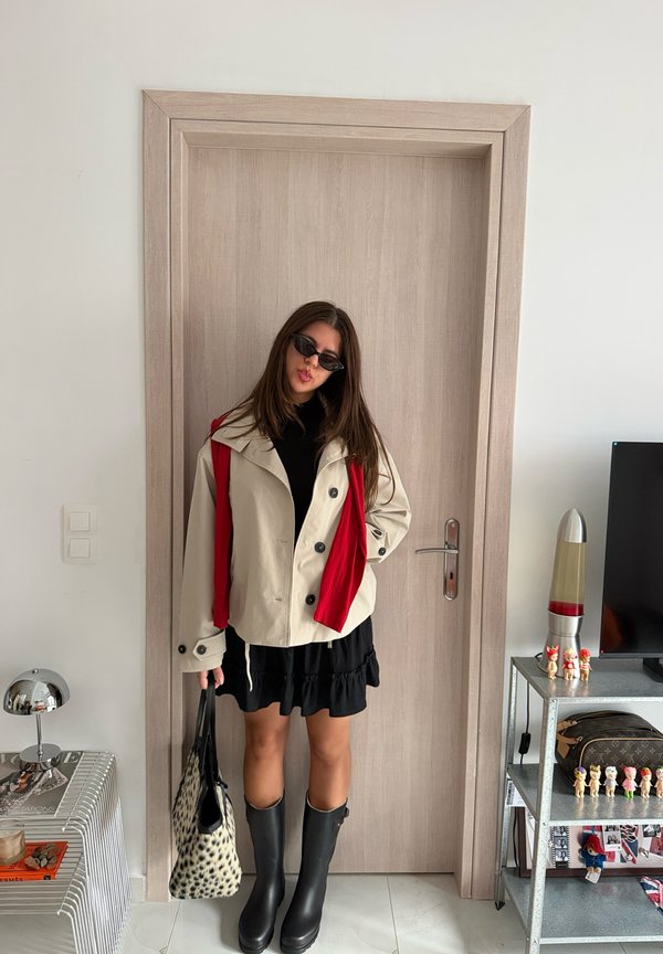 Une femme portant des lunettes de soleil, une veste crème, une jupe noire et des bottes montant jusqu’aux genoux pose près d’une porte, un sac fourre-tout à motifs à la main.