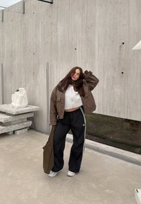 Brązowa, skórzana, oversize'owa kurtka na szarą, krótką bluzę, w połączeniu z czarnymi spodniami dresowymi z białymi paskami, trzymającą brązową, teksturowaną torbę.