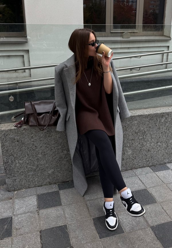 Manteau long gris sur une tunique côtelée marron, leggings noirs et baskets noir et blanc. Tenant une tasse de café, assis sur une surface en granite.