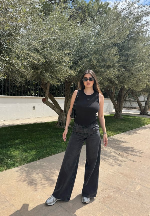 Joven mujer con camiseta negra sin mangas, vaqueros negros de pierna ancha, zapatillas deportivas y gafas de sol, de pie en una acera soleada cerca de árboles y césped.